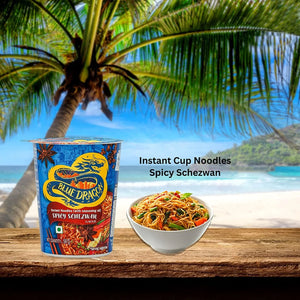 Blue Dragon Spicy Schezwan instant cup noodles and bowl of spicy noodles on wooden table with tropical beach background
