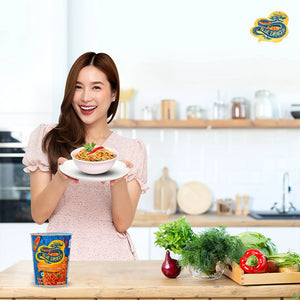 Smiling woman in kitchen holding bowl of Blue Dragon Sriracha Chilli instant noodles with fresh vegetables on table