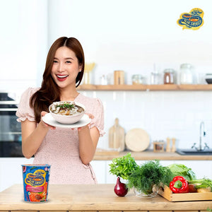 Smiling woman holding bowl of Thai sweet chilli noodles in bright kitchen with fresh herbs and Blue Dragon cup