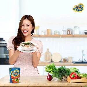 Smiling woman holding a bowl of Thai Tom Yum noodle soup in a kitchen with fresh vegetables and Blue Dragon noodles