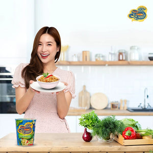 Smiling woman holding bowl of Thai green curry noodles in modern kitchen with fresh vegetables
