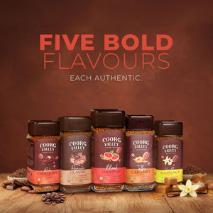 Five jars of Coorg Valley instant coffee in assorted flavors with beans and flavor ingredients on a wooden surface.