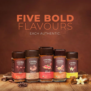 Five Coorg Valley instant coffee jars in assorted flavors on a wooden table, with beans and ingredients.