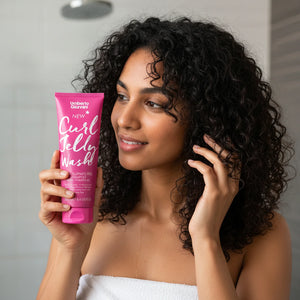 Woman with curly hair holding Umberto Giannini Curl Jelly Wash shampoo in a bathroom setting