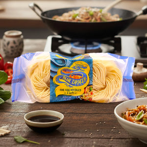Blue Dragon fine egg noodles pack on wooden table with cooked noodles in wok and vegetable stir-fry bowl