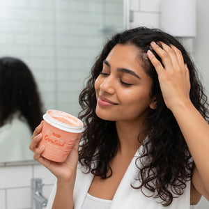 Woman with curly hair applying GrowScrub scalp scrub in bathroom with white tiles and mirror