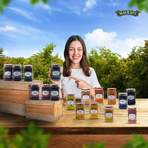Smiling woman in white holding Mackays blueberry and blackcurrant preserve jar outdoors with various preserves on wooden crates