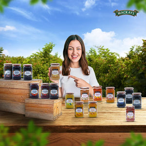 Smiling woman outdoors holding a jar of Mackays orange lemon ginger marmalade, surrounded by assorted fruit preserves on wooden table.