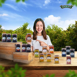 Smiling woman in white pointing to Mackays Pink Grapefruit Marmalade jar with various Mackays fruit preserves on wooden table outdoors