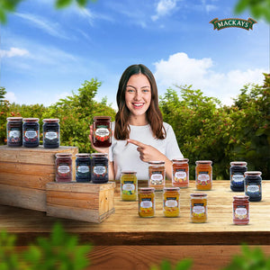 Smiling woman holding and pointing to Mackays Scottish Raspberry Preserve jar with various Mackays fruit preserves on wooden crates outdoors