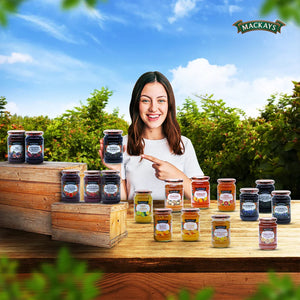 Smiling woman holding Mackays Seedless Blackberry Preserve jar outdoors with variety of Mackays preserves on wooden table