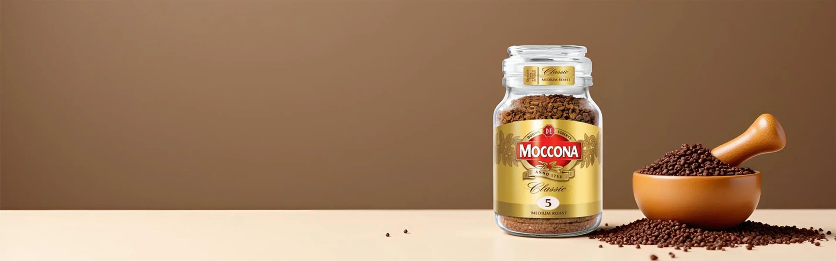 Jar of Moccona Classic Medium Roast instant coffee beside wooden bowl and scoop with coffee granules