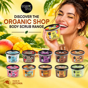 A woman smiling while holding an Organic Shop Mango Sugar Body Scrub, surrounded by various body scrub products and tropical fruits.