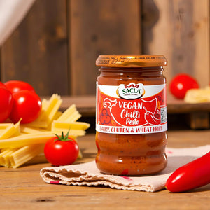Jar of Sacla vegan chilli pesto on cloth with tomatoes, red chili, and pasta on wooden table