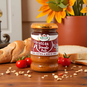 Jar of Sacla vegan tomato pesto on wooden table with cherry tomatoes, bread, nuts, and sunflower in orange pot