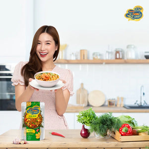 Smiling woman in kitchen holding bowl of shrimp chow mein with Blue Dragon noodle kit and fresh vegetables on counter