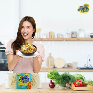 Smiling woman holding a bowl of Pad Thai noodles in a bright kitchen with Blue Dragon rice noodles and fresh vegetables