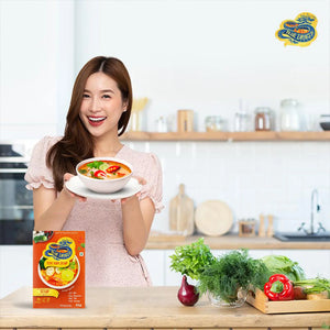 Smiling woman in modern kitchen holding bowl of tom yum soup with Blue Dragon soup kit and fresh vegetables