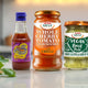 Assorted gourmet sauces including sweet chili, whole cherry tomato pasta sauce, and vegan basil pesto on kitchen counter with fresh vegetables