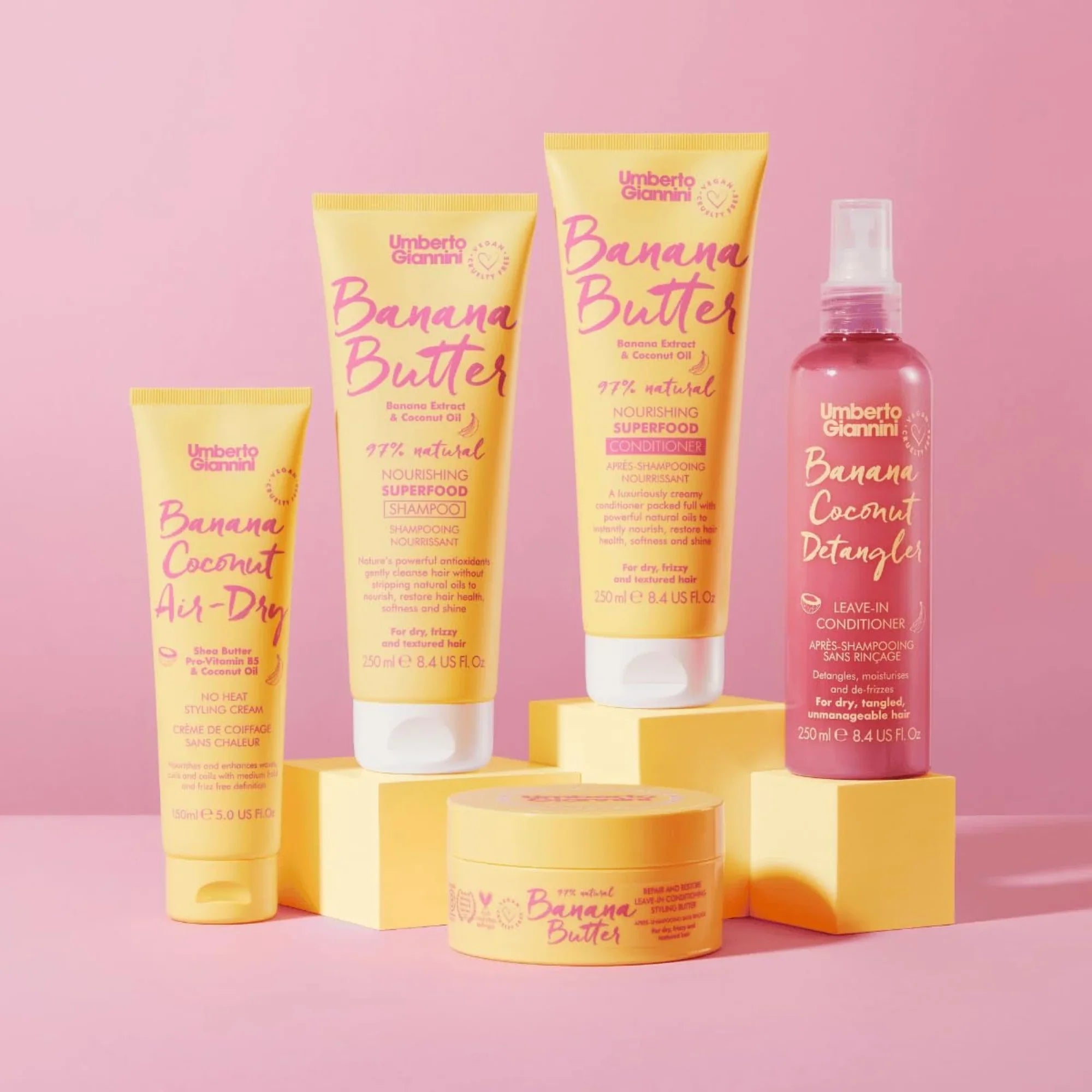 Umberto Giannini Banana Butter and Coconut haircare products including shampoo, conditioner, detangler, and styling cream on pink background