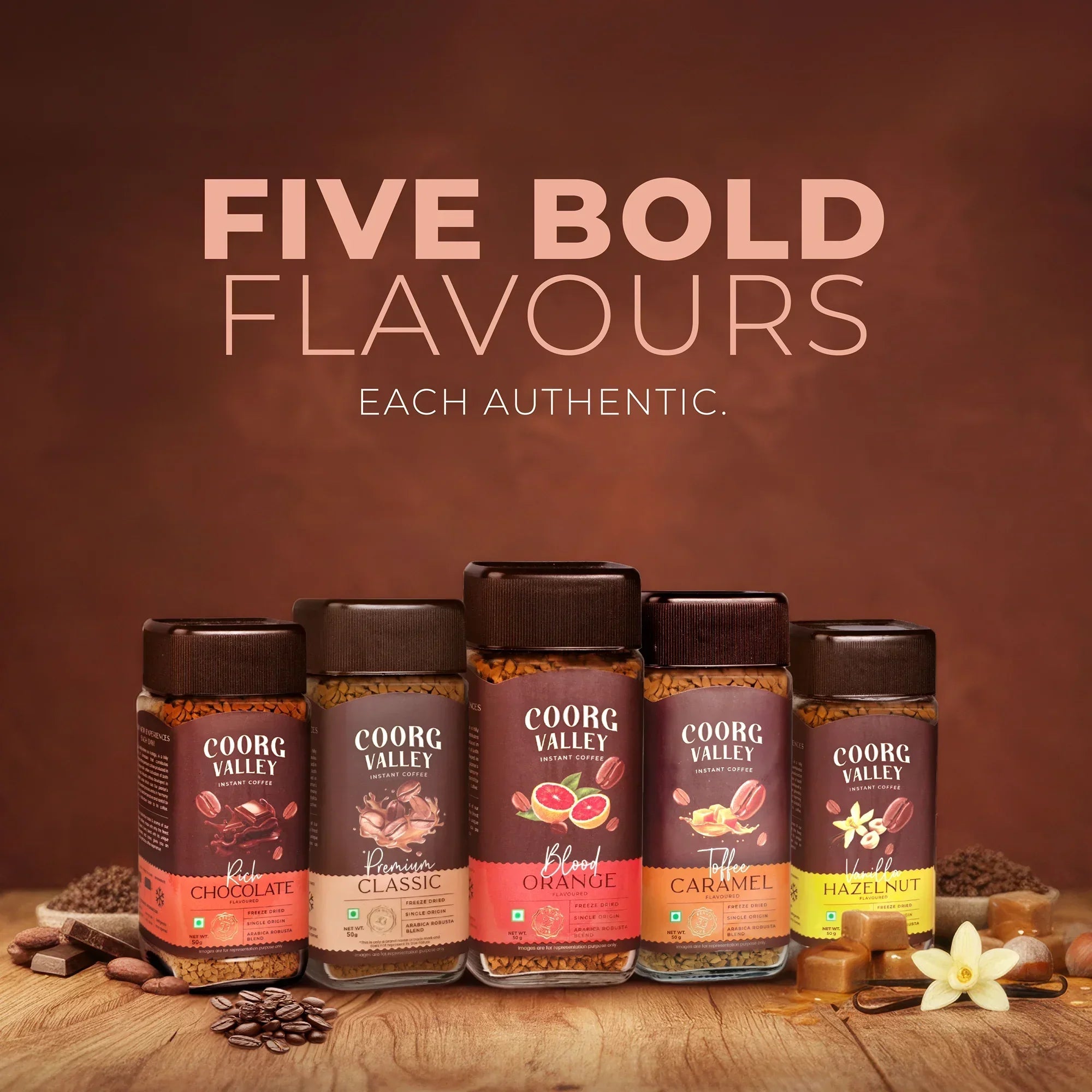 Five jars of Coorg Valley instant coffee in chocolate, classic, orange, caramel, and hazelnut flavors on a wooden surface with matching ingredients.