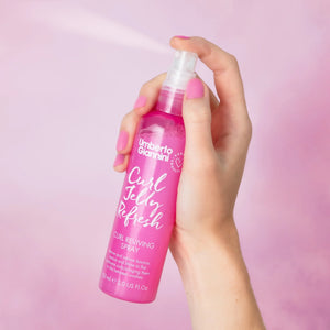 Hand holding Umberto Giannini Curl Jelly Refresh curl reviving spray in pink bottle with light pink background