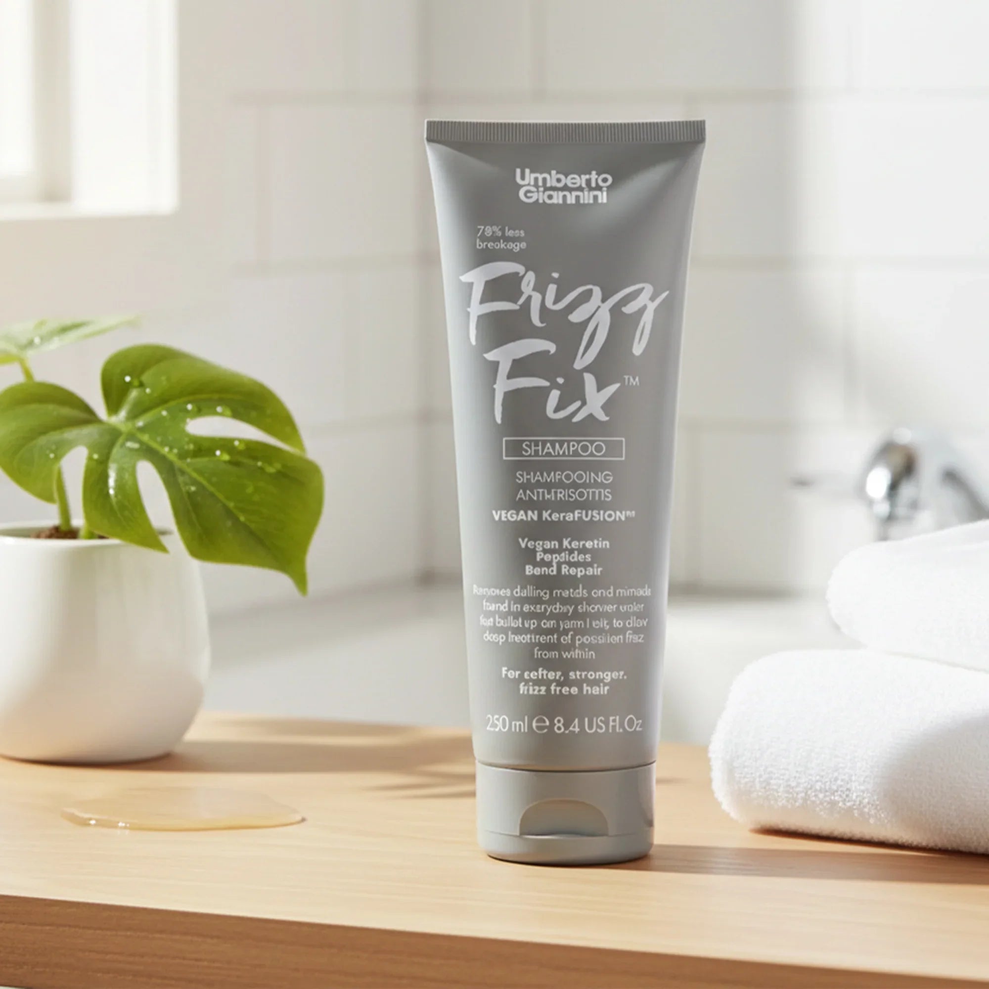 Umberto Giannini Frizz Fix Vegan Keratin Shampoo tube on wooden surface with plant and white towels