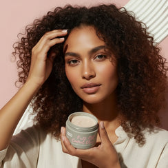 Woman with curly hair holding a jar of Fuzz-Off Control Clay hair product against a blush pink background