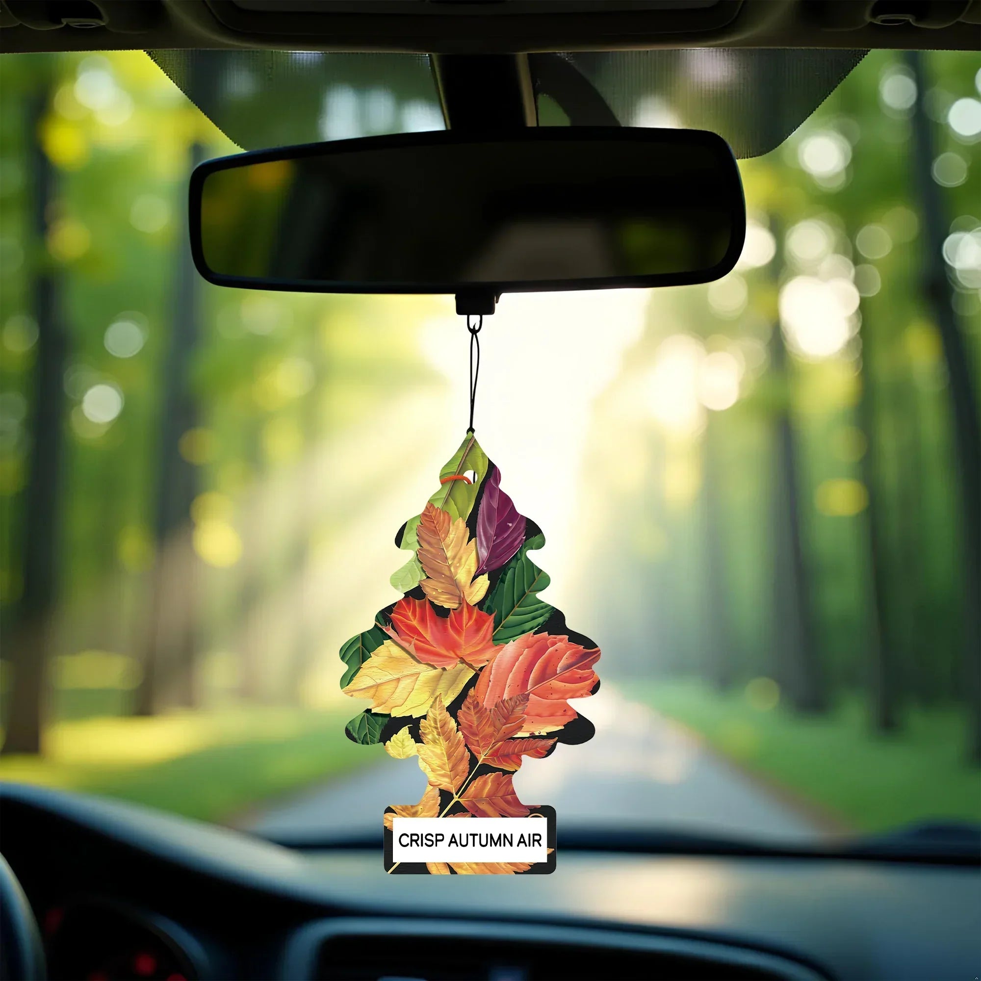 Car air freshener with autumn leaves hanging from a car's rearview mirror, with a forest view outside.