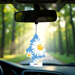 Daisy Fields Little Trees car air freshener hanging from rearview mirror with sunlight and forest road background