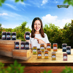 Smiling woman pointing to Mackays blueberry preserve jar with various jars of Mackays fruit preserves on wooden table outdoors