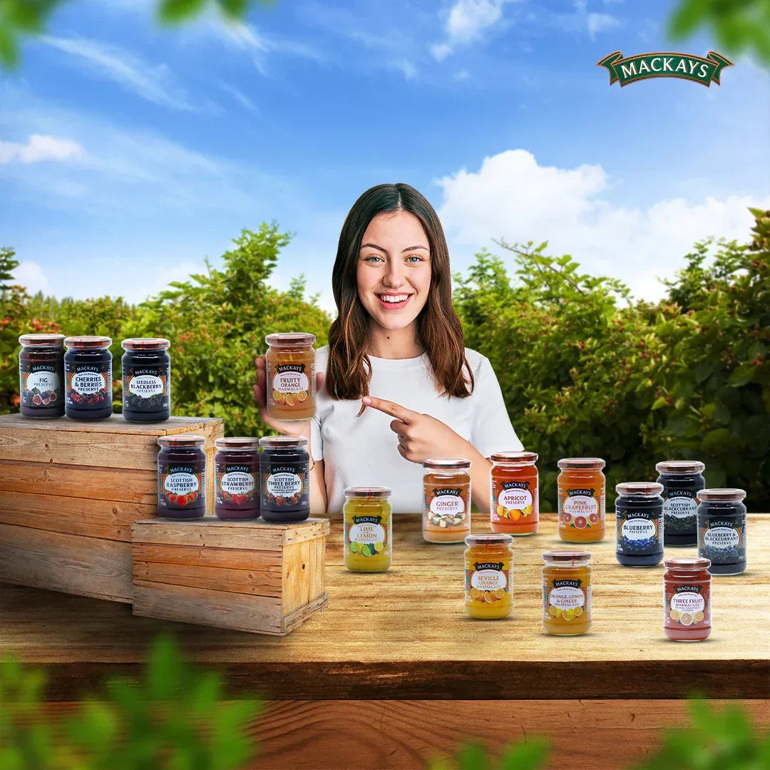 Woman smiling and pointing at Mackays fruity orange marmalade jar surrounded by various Mackays fruit preserves on wooden table outdoors