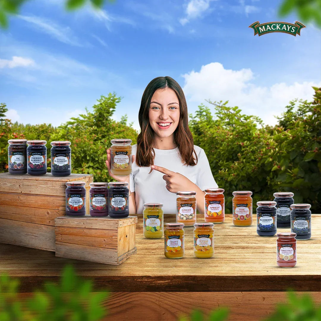 Smiling woman holding Mackays orange lemon ginger marmalade jar with various Mackays preserves jars on wooden table outdoors
