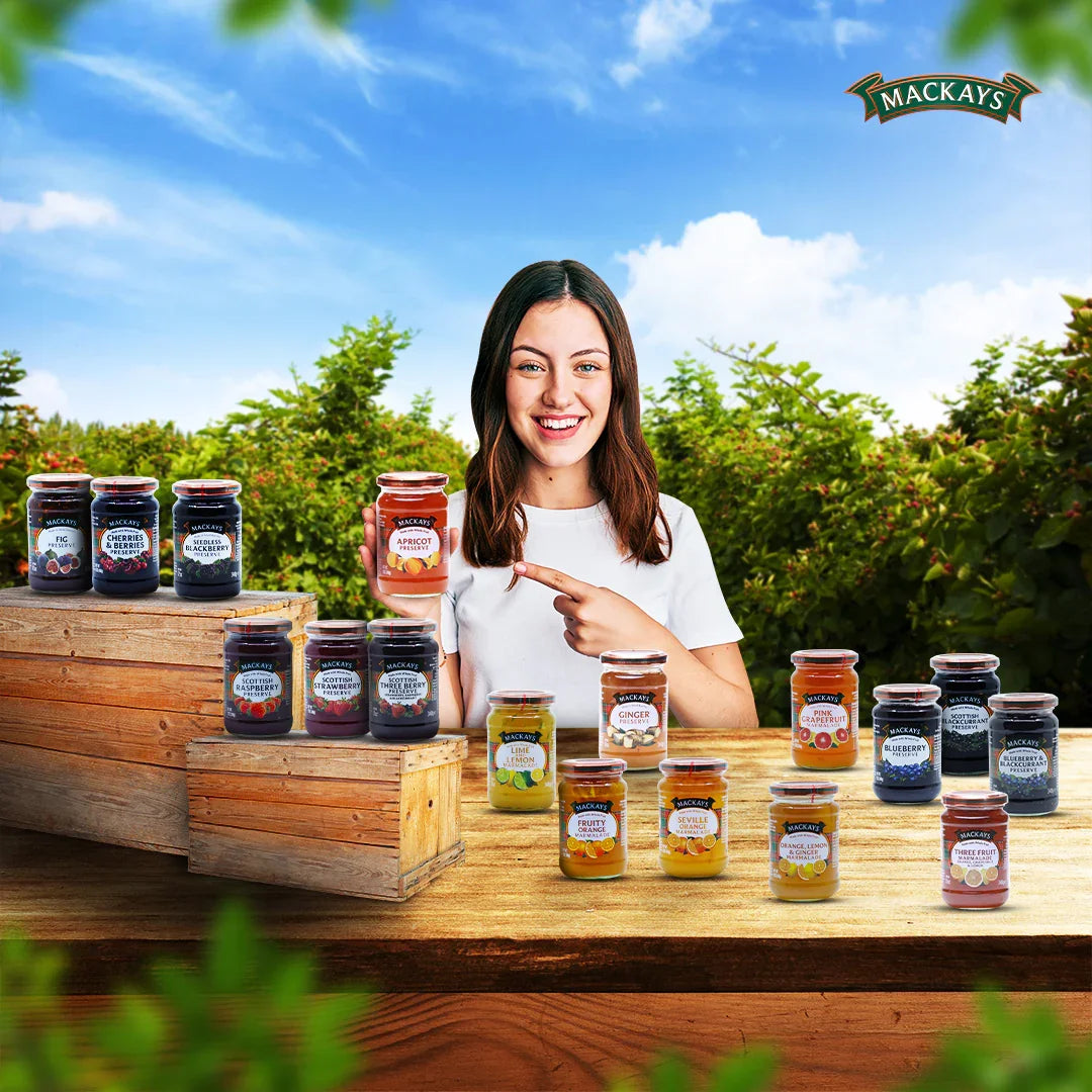Smiling woman holding Mackays apricot preserve jar surrounded by various Mackays fruit preserves on a rustic wooden table with green foliage background