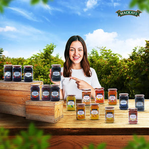 Smiling woman in white holds Mackays Scottish Blackcurrant Preserve jar surrounded by various Mackays fruit preserves outdoors