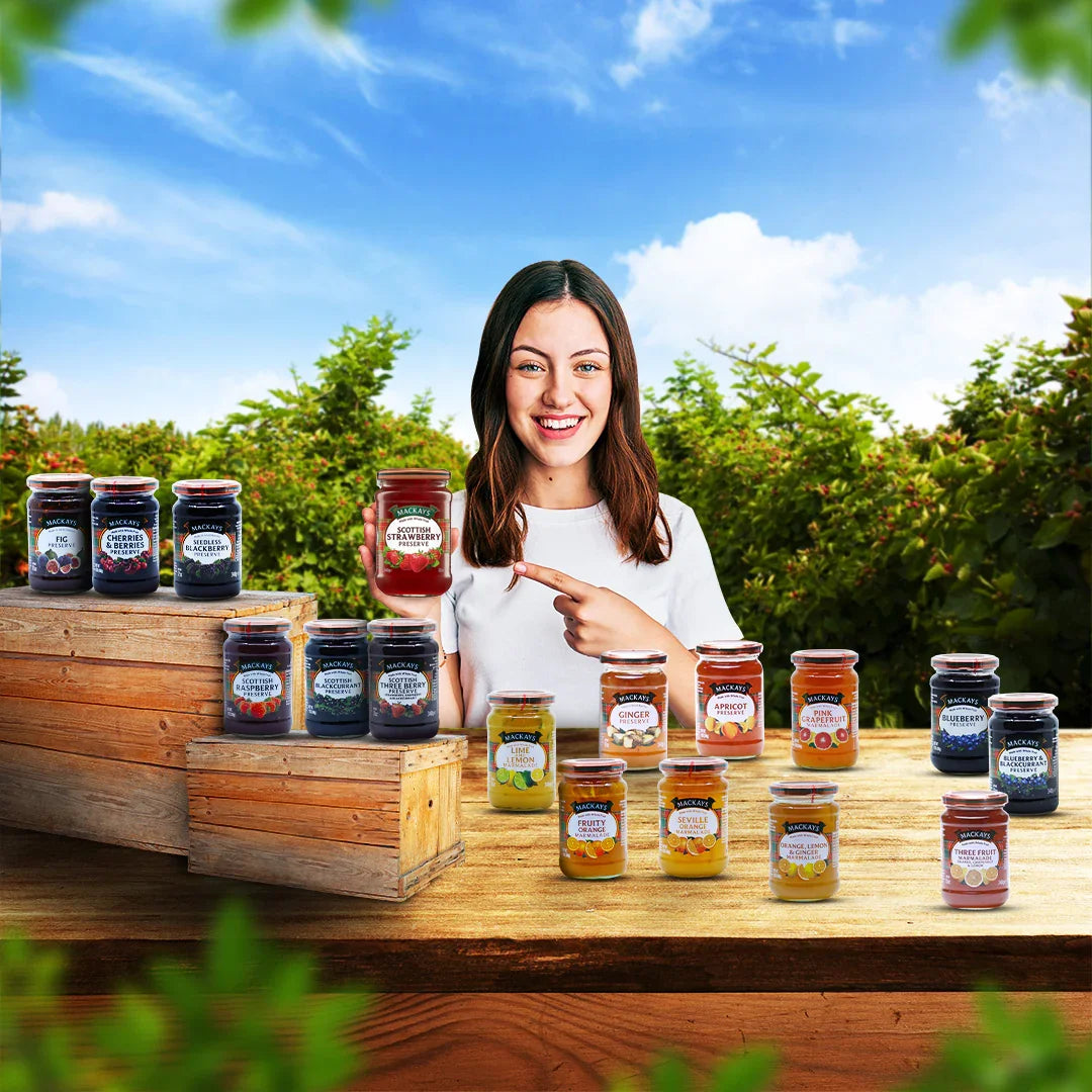 Smiling woman outdoors holding Mackays Scottish Strawberry Preserve with assorted fruit jam jars on wooden crates