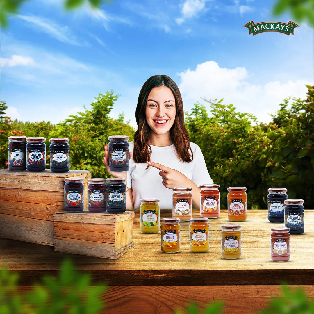 Woman outdoors holding Mackays Scottish Three Berry Preserve jar, surrounded by assorted fruit preserves on wooden crates