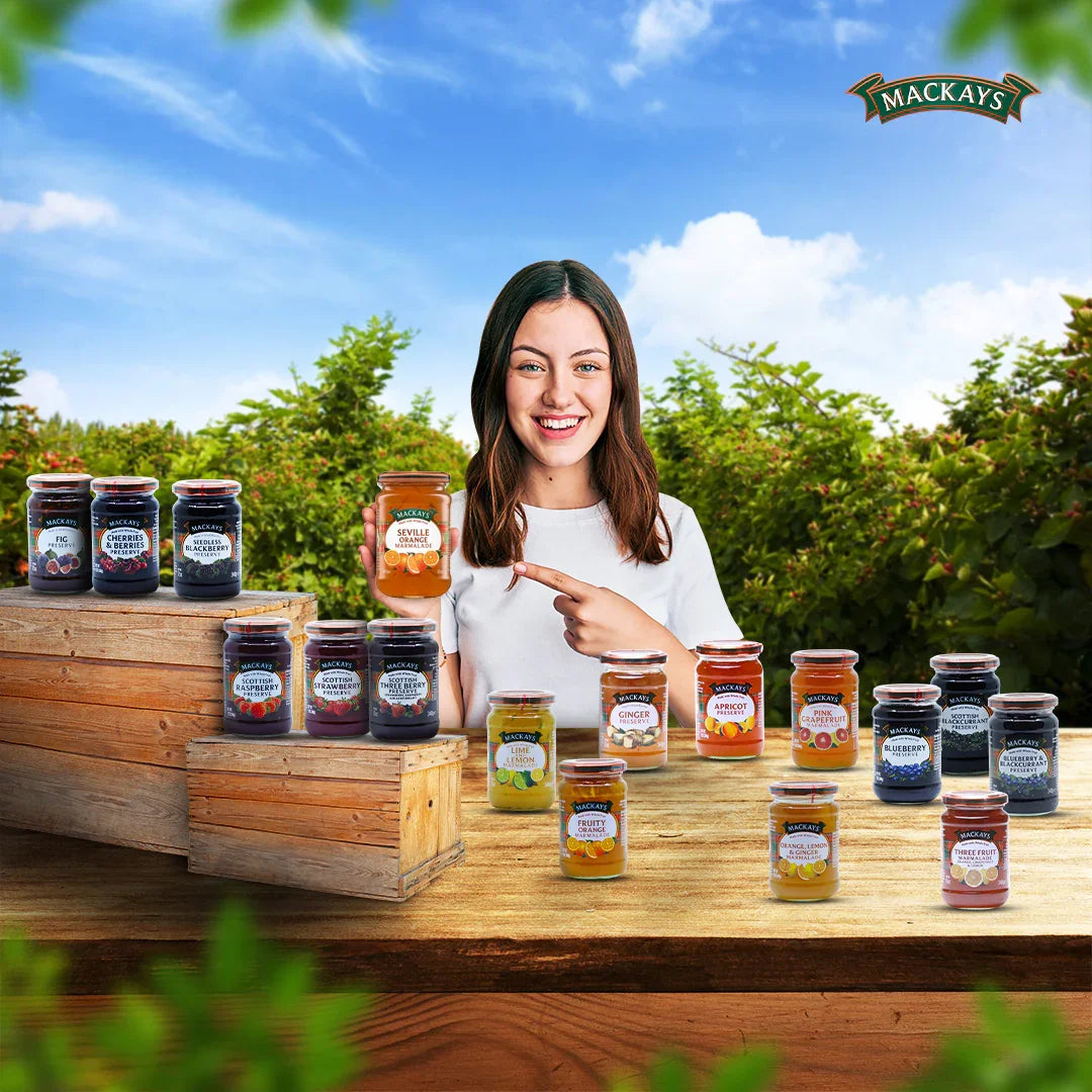 Smiling woman holds Mackays Seville Orange Marmalade jar with various Mackays jams and marmalades on wooden table outdoors