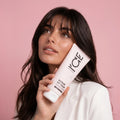 Woman holding a tube of Repair My Hair hair mask against a pink background.