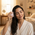 Woman in white robe brushing wet hair with pink scalp massager in cozy bedroom
