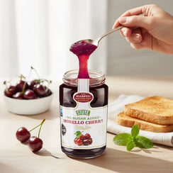 Jar of Stute No Sugar Added Morello Cherry jam with spoonful of jam, fresh cherries in bowl, toast, and mint on wooden table