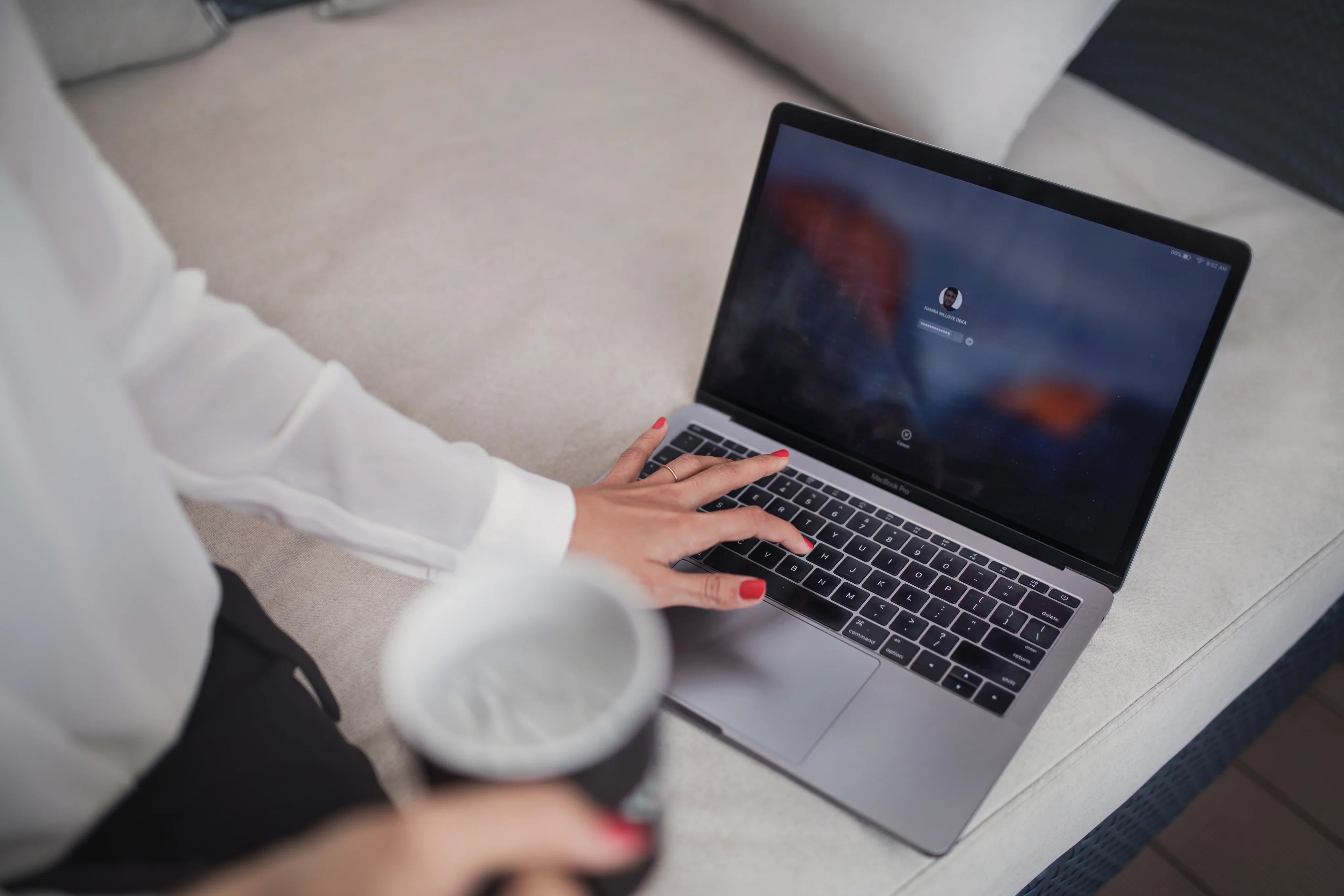 Person with red nail polish logging into MacBook Pro laptop on beige couch while holding a coffee cup