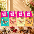 Four boxes of Pikk Pop Waffle Wonders mini cookies in assorted flavors, arranged on a table with bowls of vanilla ice cream, raspberries, caramel, almonds, and chocolate, against a warm kitchen background.