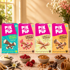 Four boxes of Pikk Pop Waffle Wonders mini cookies in assorted flavors, arranged on a table with bowls of vanilla ice cream, raspberries, caramel, almonds, and chocolate, against a warm kitchen background.